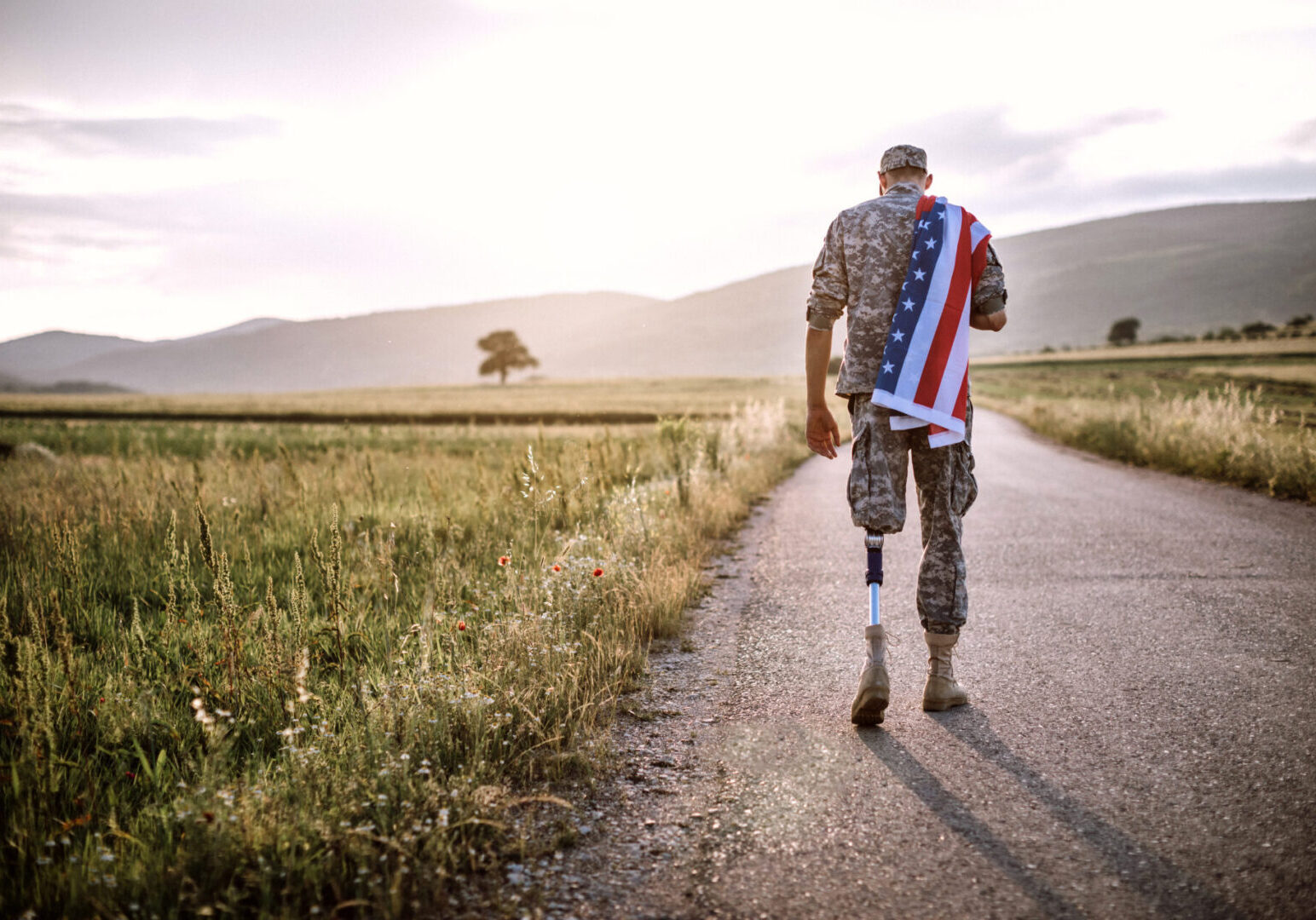 Rear View Of Young Amputee Soldier Walking Road Wearing American Flag, unrecognizable person