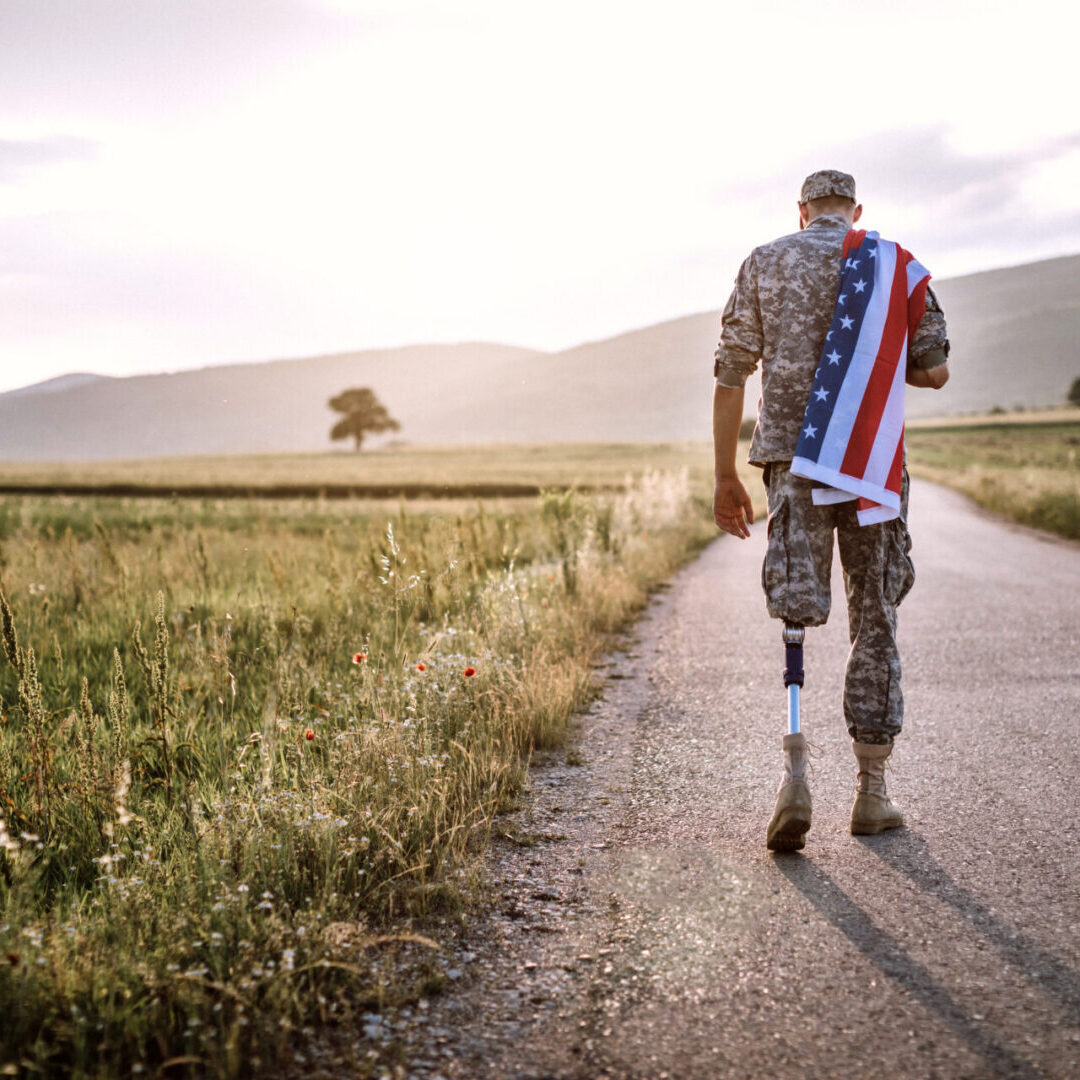 Rear View Of Young Amputee Soldier Walking Road Wearing American Flag, unrecognizable person